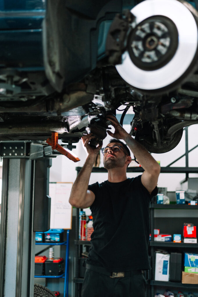 Auto repair technician using power tools under a lifted car in a body shop